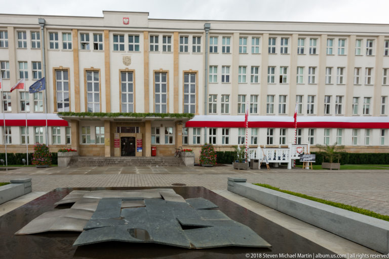 Symmetrical multi-story institutional building facing a paved square with low dark metal sculptural plates in the foreground.
