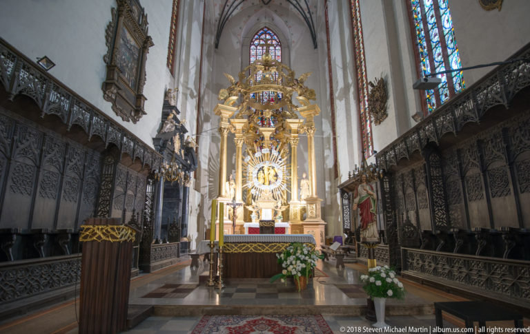 Gilded high altar centered between dark carved choir stalls, with tall stained-glass windows in a Gothic church sanctuary.