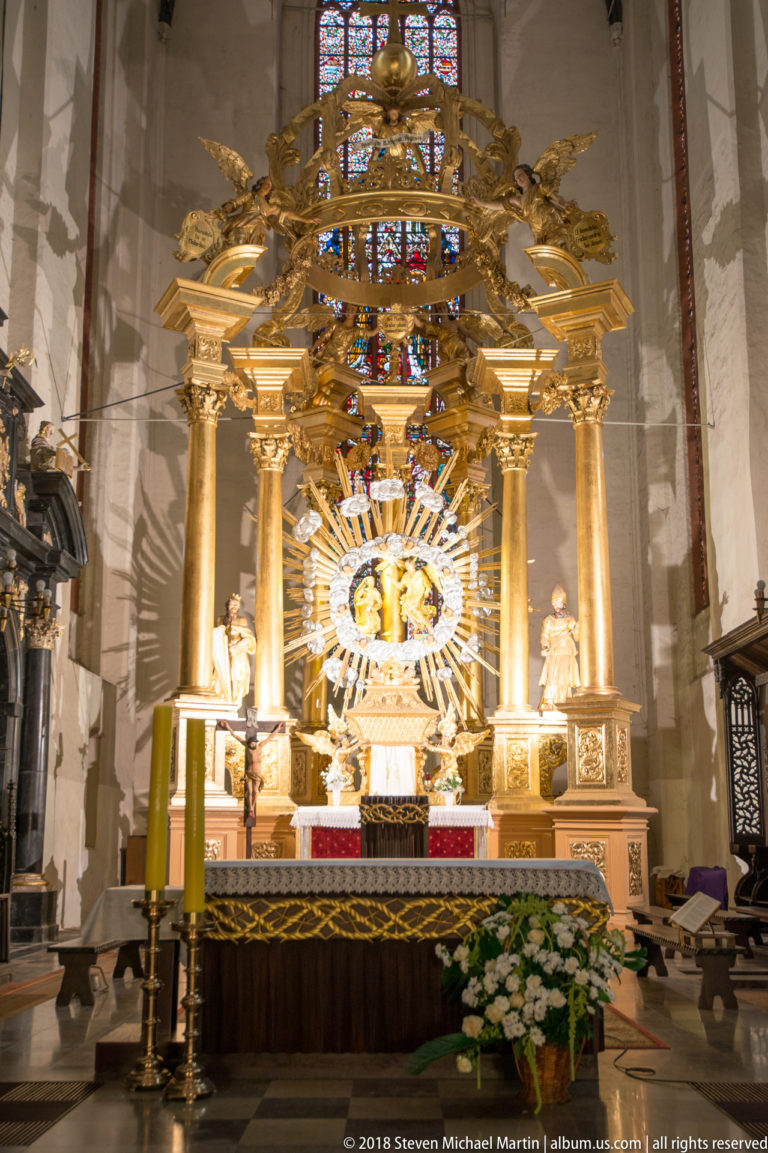 Large gilded Baroque high altar with columns, sculpted figures, radiant central ornament, flowers, and stained glass behind.