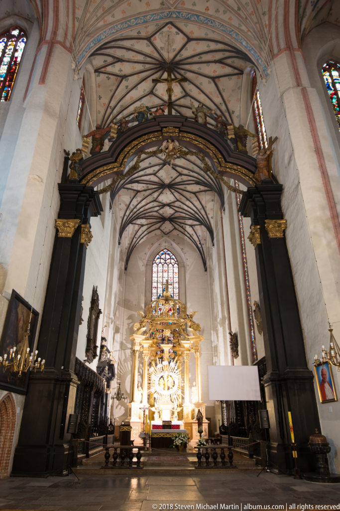 Gothic church nave with painted ribbed vaults and a dark wooden screen framing a distant gilded high altar.