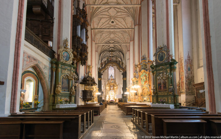 Long Gothic church interior with rows of wooden pews, green-and-gold side altars, chandeliers, and a bright sanctuary in the distance.
