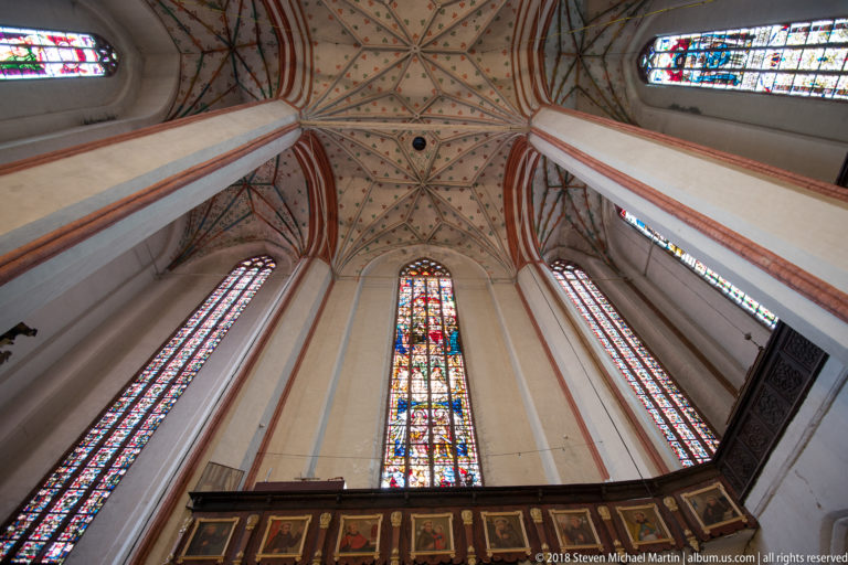 Upward view of painted Gothic rib vaults and tall stained-glass windows above dark carved church furnishings.