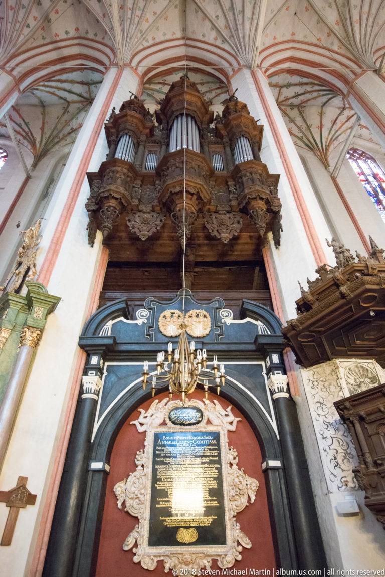 Historic pipe organ on a carved loft above a large black-and-white inscribed memorial panel in a Gothic church.
