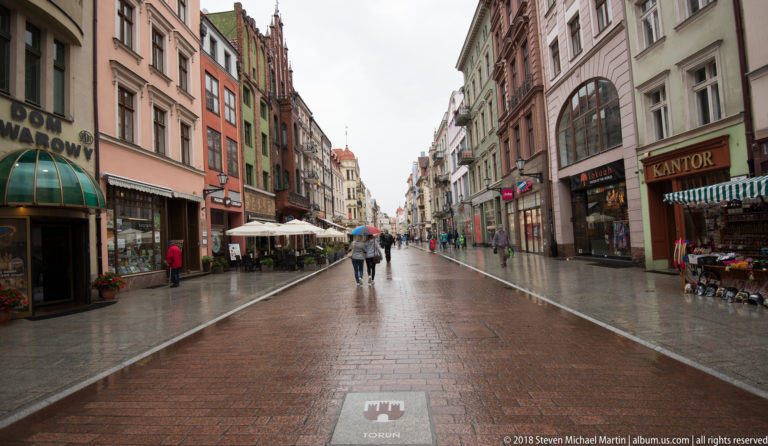 Rainy pedestrian street lined with colorful historic buildings, shops, café seating, and people carrying umbrellas in Toruń Old Town.