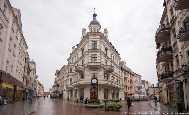 Pale historic corner building with columns, balconies, a rooftop tower, and a large projecting clock at a Toruń street junction.