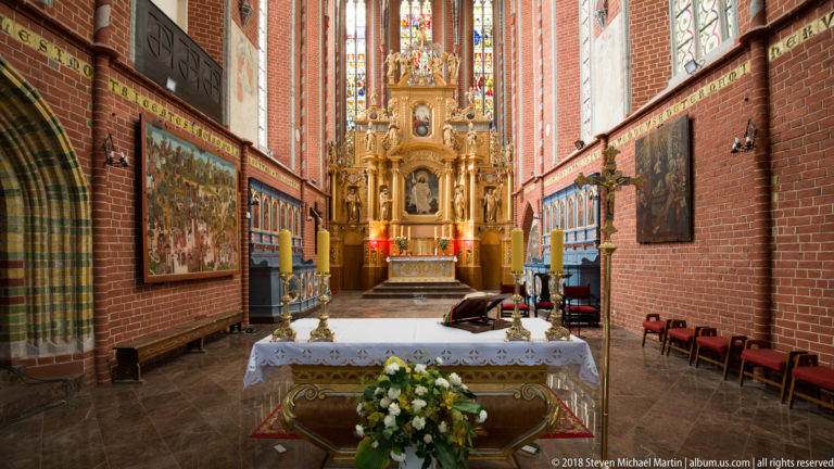 Red-brick Gothic sanctuary with tall stained-glass windows, a gilded altar, side paintings, white altar cloth, and flowers.