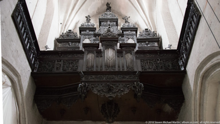 Historic pipe organ in an ornate carved wooden case and loft inside a Gothic church.