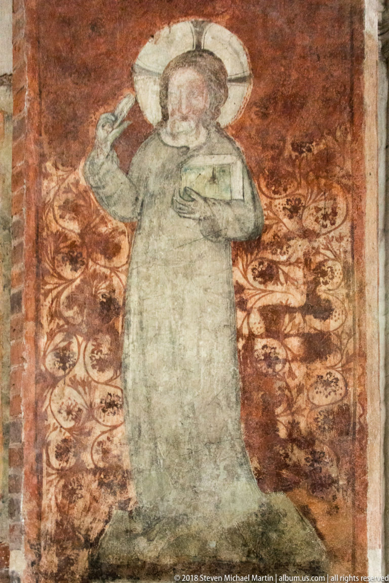 Faded medieval wall painting of a haloed saint against a patterned red background.