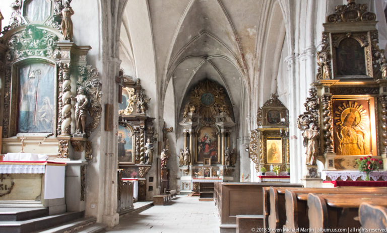 Gothic church side aisle lined with altars, sculptures and devotional images beneath pointed vaults.