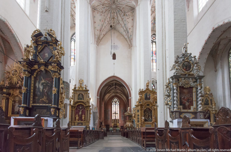 Tall Gothic nave with white piers, pointed arches, carved Baroque altars, wooden pews, and a distant stained-glass window.