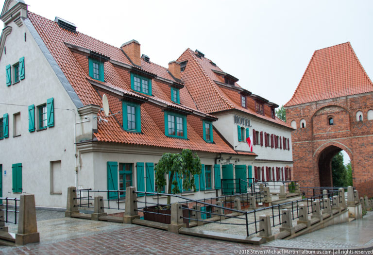 Historic hotel buildings with green shutters beside a large brick medieval gate in Toruń.