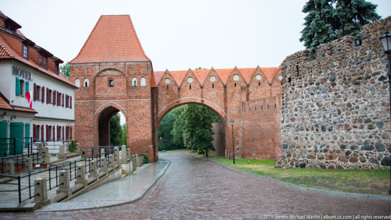 Brick medieval city gate and adjoining fortified wall spanning a cobbled street in Toruń.