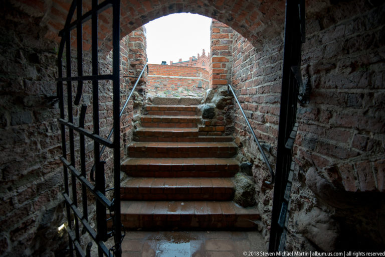 Brick stairway with iron railings ascending through a narrow vaulted passage in castle ruins.