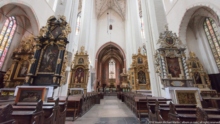 Wide nave view with large gilded side altars, pointed Gothic arches, stained-glass windows, and rows of wooden seating.