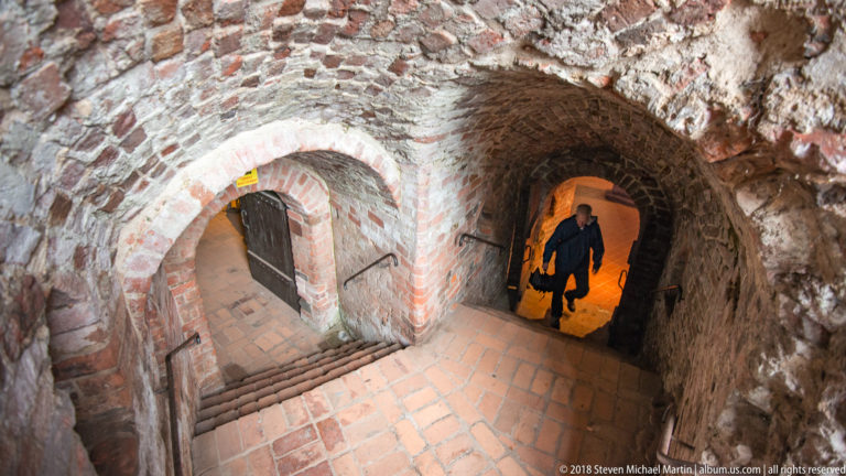 Intersecting brick stairways and vaulted passages inside the Teutonic Castle ruins, with a visitor at right.