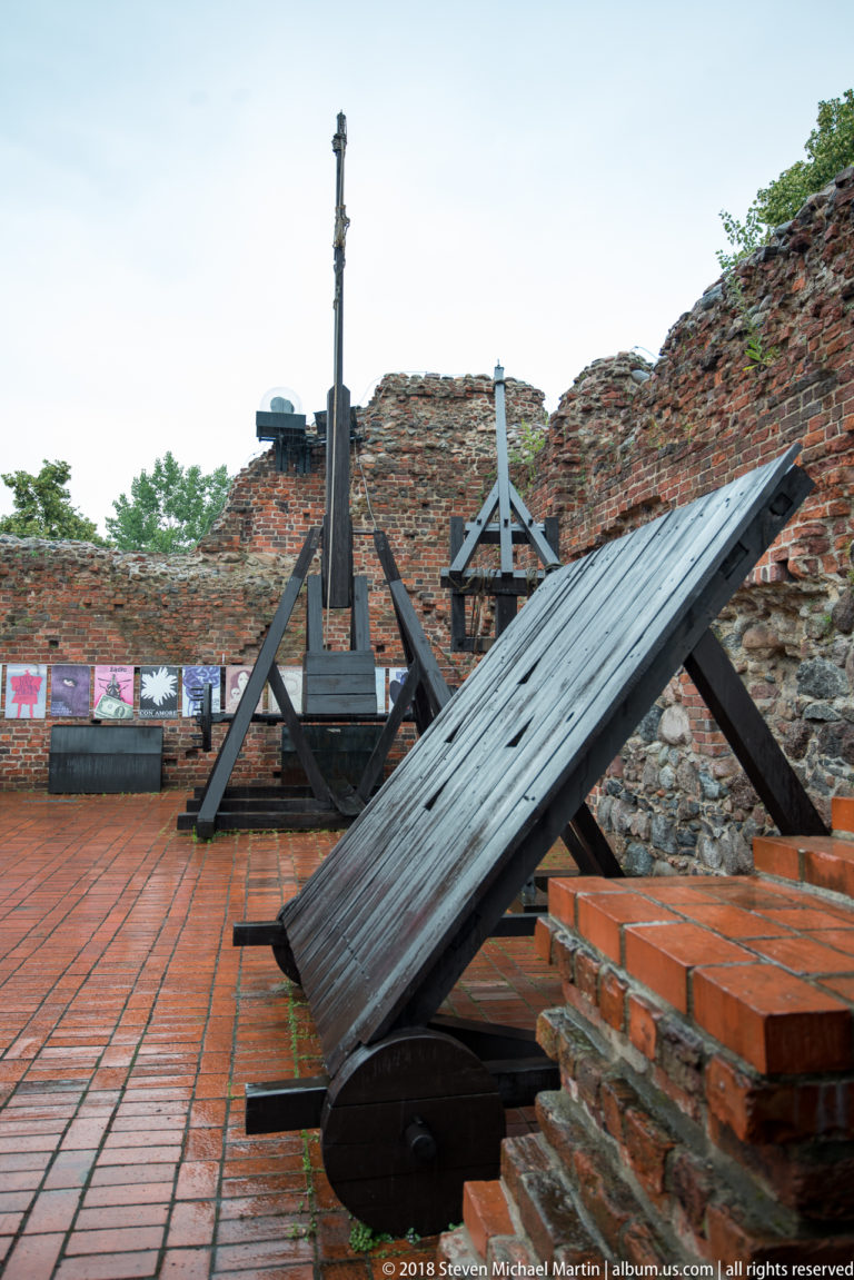 Wooden replicas of medieval siege engines displayed on brick paving inside castle ruins.