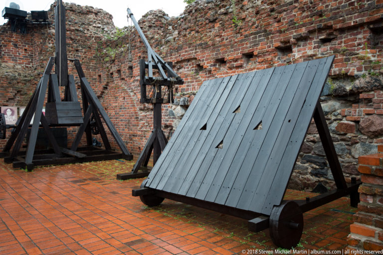 Large wooden siege-engine reconstruction beside weathered brick walls of the Teutonic Castle ruins.