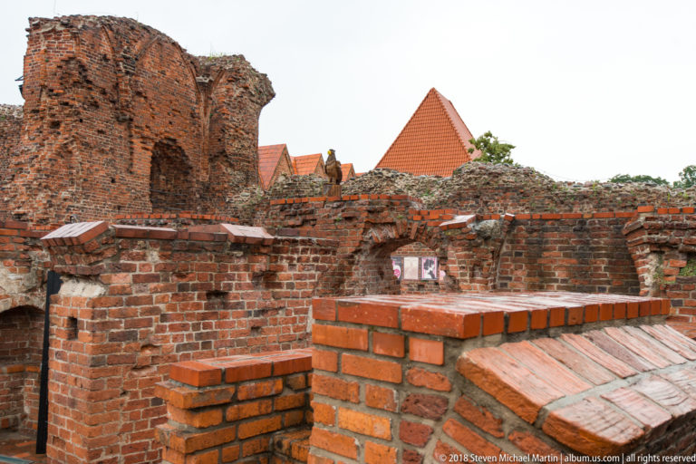 Fragmented brick walls and foundations of the Teutonic Castle ruins with city roofs beyond.