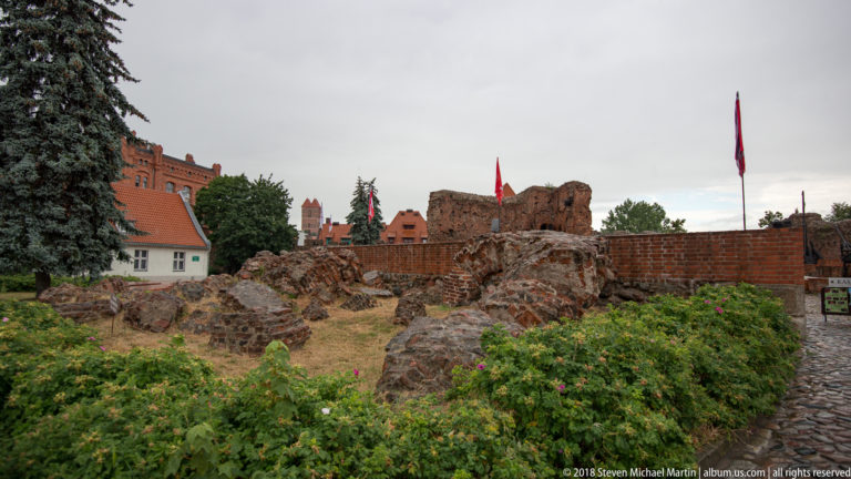 Open-air stone and brick ruins of the Teutonic Castle with walls, roofs and flags beyond.