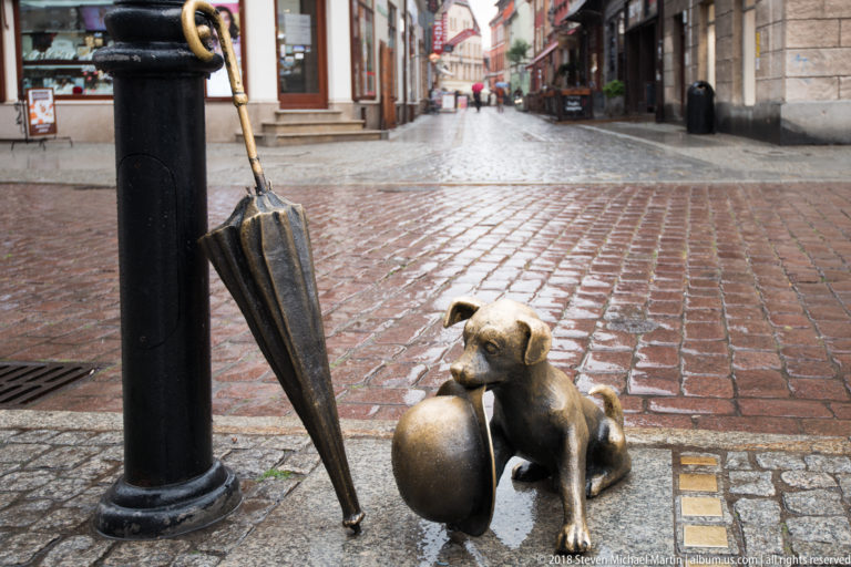 Bronze Filuś dog sculpture beside an umbrella and bowler hat on a cobbled street in Toruń.