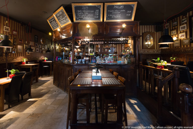 Wood-paneled pub interior with central bar, long tables, hanging lamps and framed memorabilia.