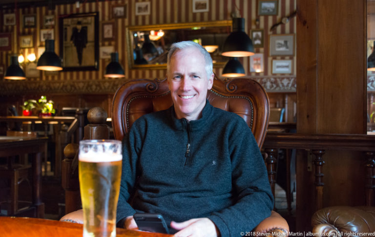 Smiling seated visitor with a glass of beer in a wood-paneled pub interior.