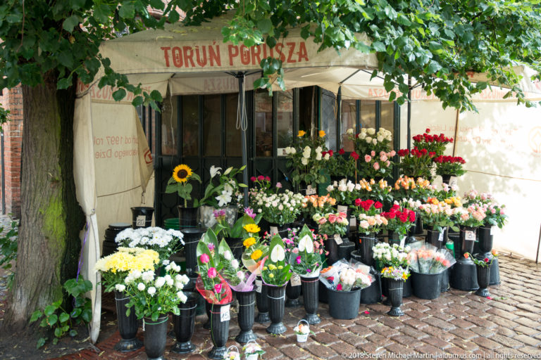 Buckets of colorful flowers and bouquets displayed outside a small shop on a cobbled Toruń street.