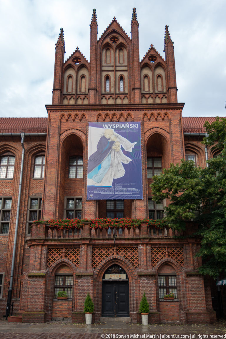 Ornate red-brick façade with stepped gables, pointed arches, balcony and large banner in Toruń.