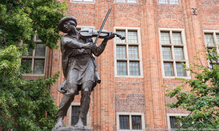 Bronze violinist sculpture playing before a red-brick historic building in Toruń.