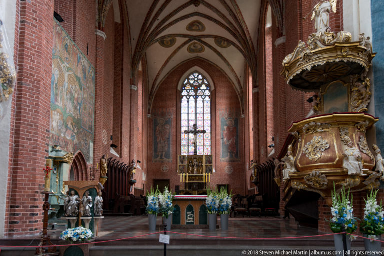 Red-brick Gothic chancel with stained glass, painted wall decoration, flowers, and a large carved Baroque pulpit at right.