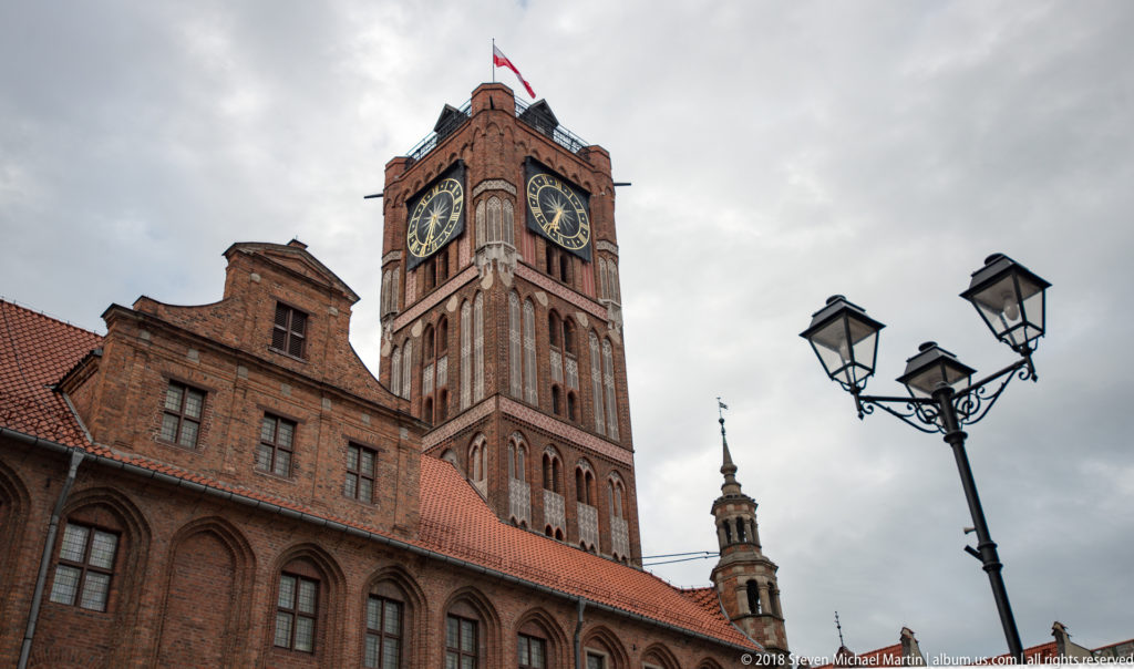 Brick clock tower of Toruń Old Town Hall with large clock faces and Polish flag.