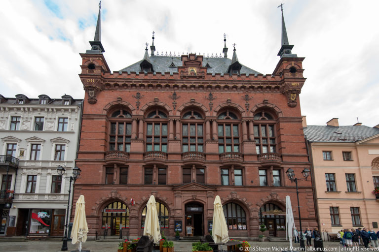 Large ornate red-brick civic building with arched windows, turrets and café seating on Toruń market square.