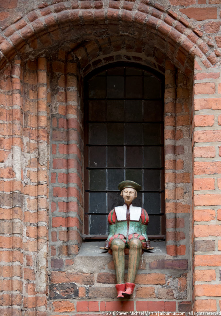 Small seated painted figurine in a barred recessed window of a red-brick historic building.