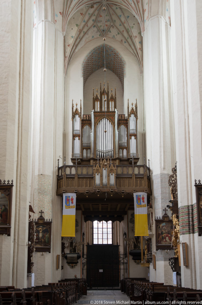 Large wooden pipe organ on a west gallery beneath a painted vault, with banners and framed religious paintings below.