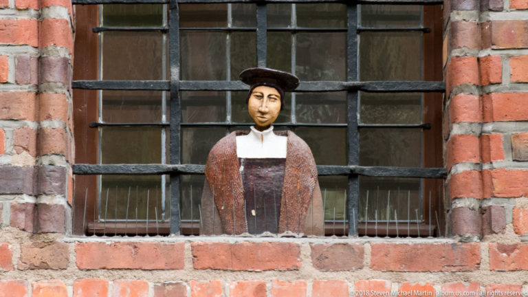 Painted standing figurine behind iron bars in a recessed red-brick window.