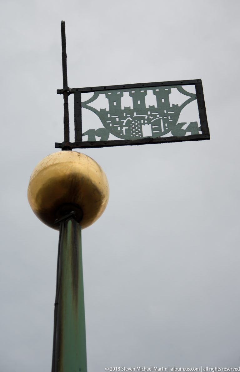 Rooftop finial supporting a cut-metal silhouette of towers and historic buildings against the sky.