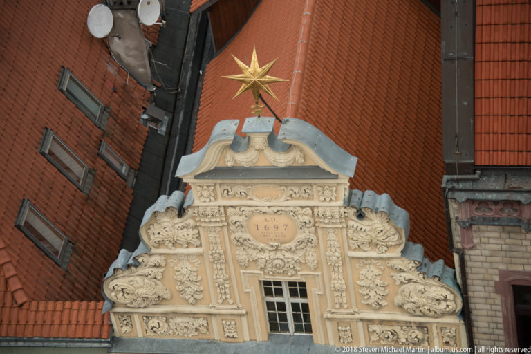 Ornate stucco gable dated 1697 with carved scrollwork and gilded star between red-tiled roofs.