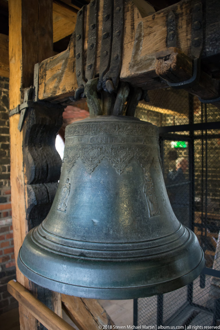 Large bronze bell suspended beneath heavy wooden beams and iron fittings in a historic belfry.
