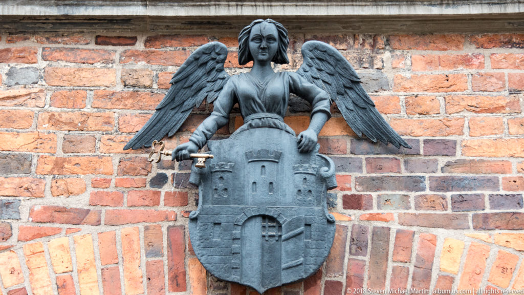 Winged female figure holding a shield with Toruń’s city coat of arms against a brick wall.