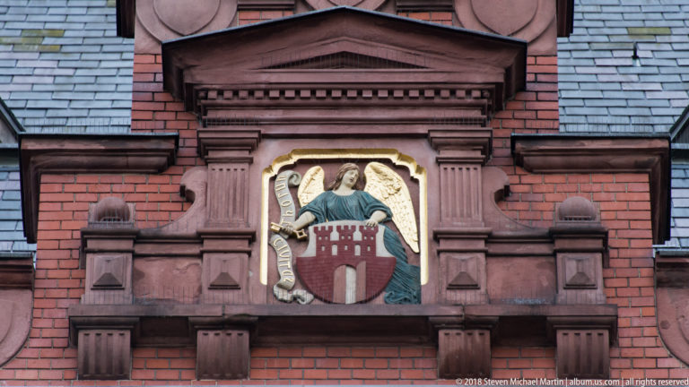 Polychrome winged figure holding Toruń’s gate-shaped coat of arms on a red-brick façade.