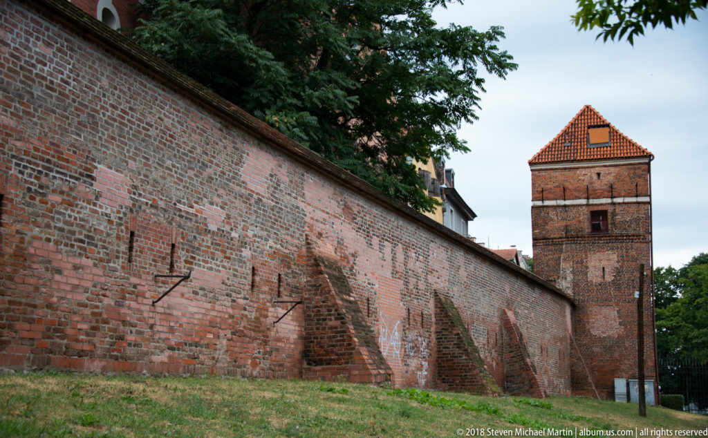 Long medieval brick city wall with buttresses leading to a square tiled-roof defensive tower.