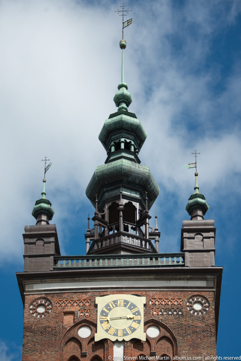 Close view of a brick church tower clock beneath a tiered green cupola and corner turrets.