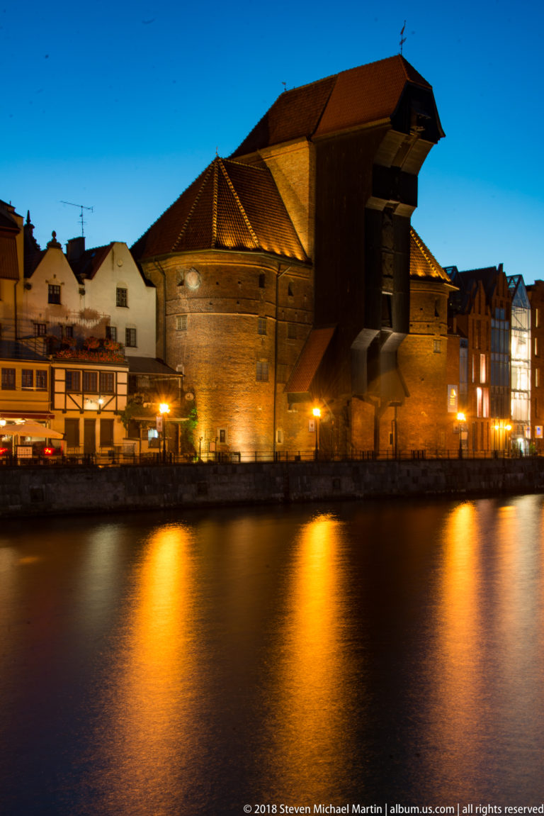 Floodlit Gdańsk Crane with brick walls, steep roofs, and dark timber hoisting structure reflected in the Motława River.