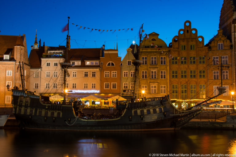 Broadside view of a dark historic-style sailing ship with masts and rigging moored before illuminated gabled houses.