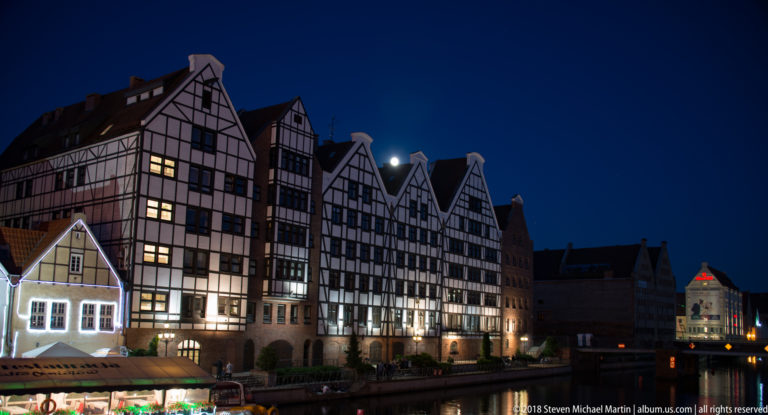 Illuminated steep-gabled half-timbered waterfront buildings reflected along a dark canal or river edge at night.