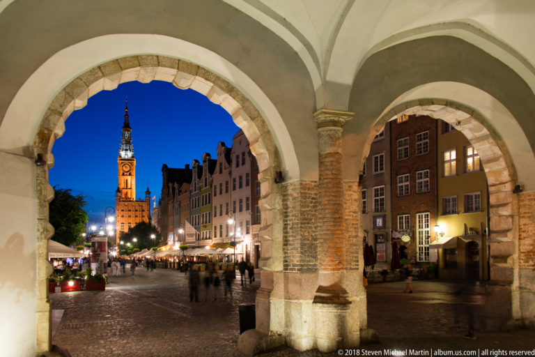 Illuminated Main Town Hall tower seen through broad masonry arches framing a cobbled pedestrian street at night.