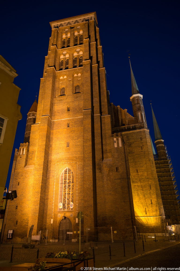 Large floodlit Brick Gothic church with heavy buttresses, tall pointed windows, and slender corner spires at night.