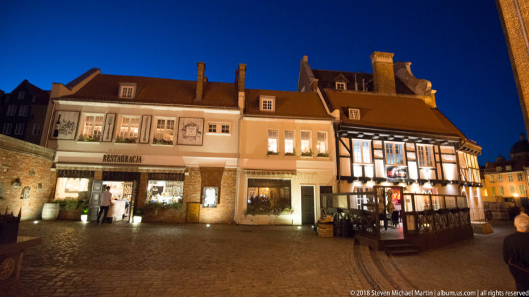 Small cobbled square with illuminated restaurant façades, a pale stucco building, and a neighboring half-timbered house.