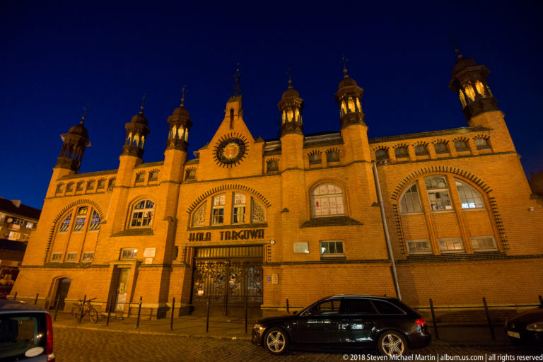 Illuminated brick Hala Targowa with large arched windows, decorative turrets, central entrance sign, parked cars, and bicycles.