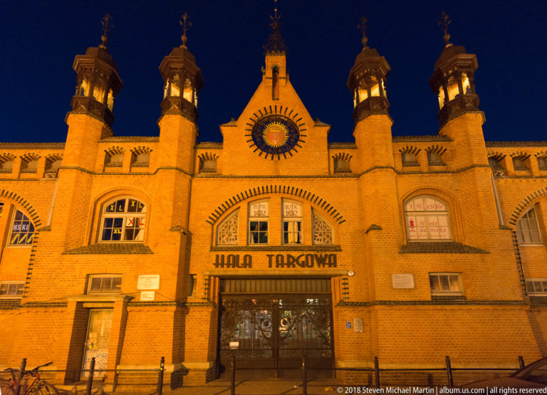 Closer nighttime view of Hala Targowa entrance with name sign, circular window, arched brickwork, and small roof turrets.